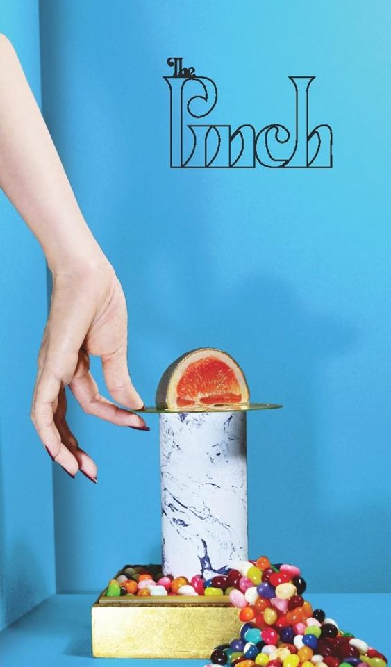 Cover of The Pinch: a hand reaching toward a grapefruit slice balanced on a marble column against a blue wall